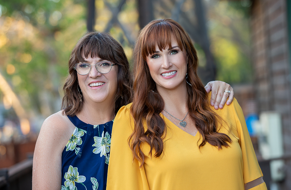 Two women standing next to each other in front of a tree.