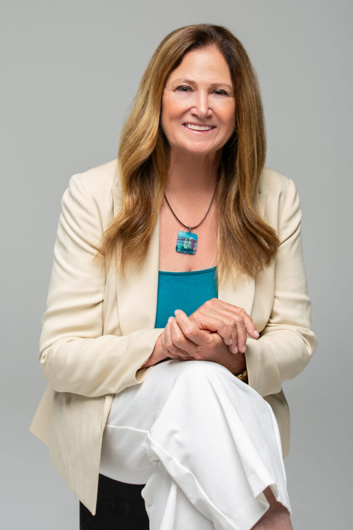 A woman in white jacket sitting on chair with hands clasped.